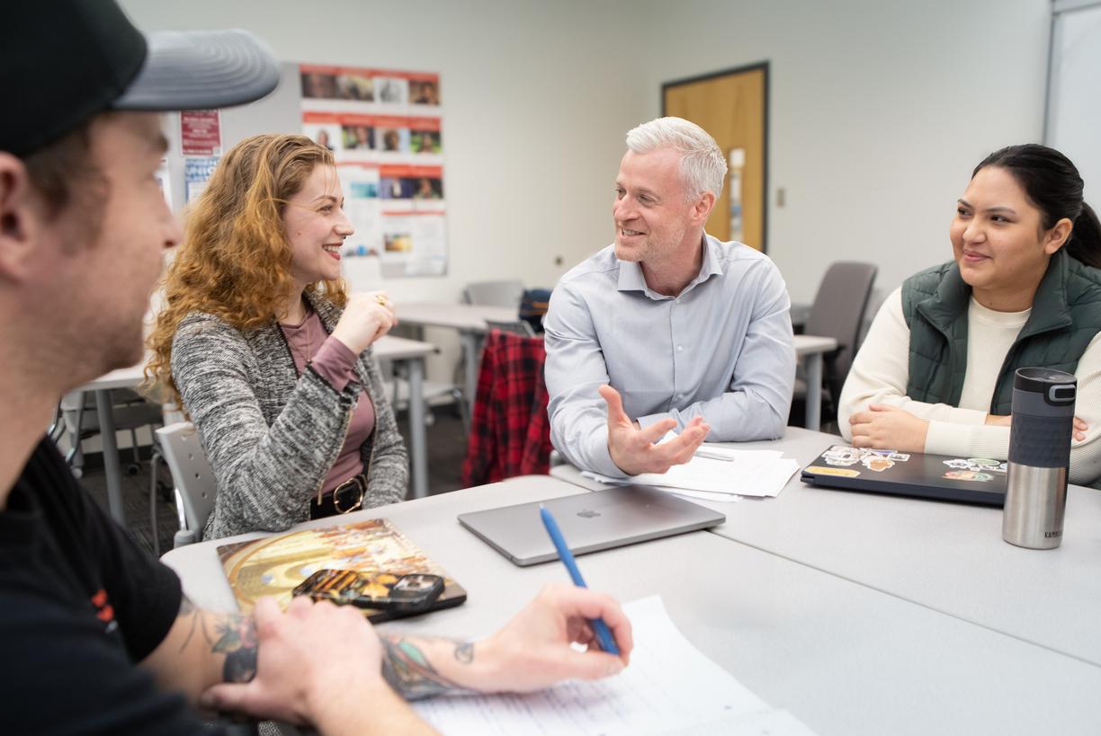 Four people talk at a table with laptops and notepads open in front of them