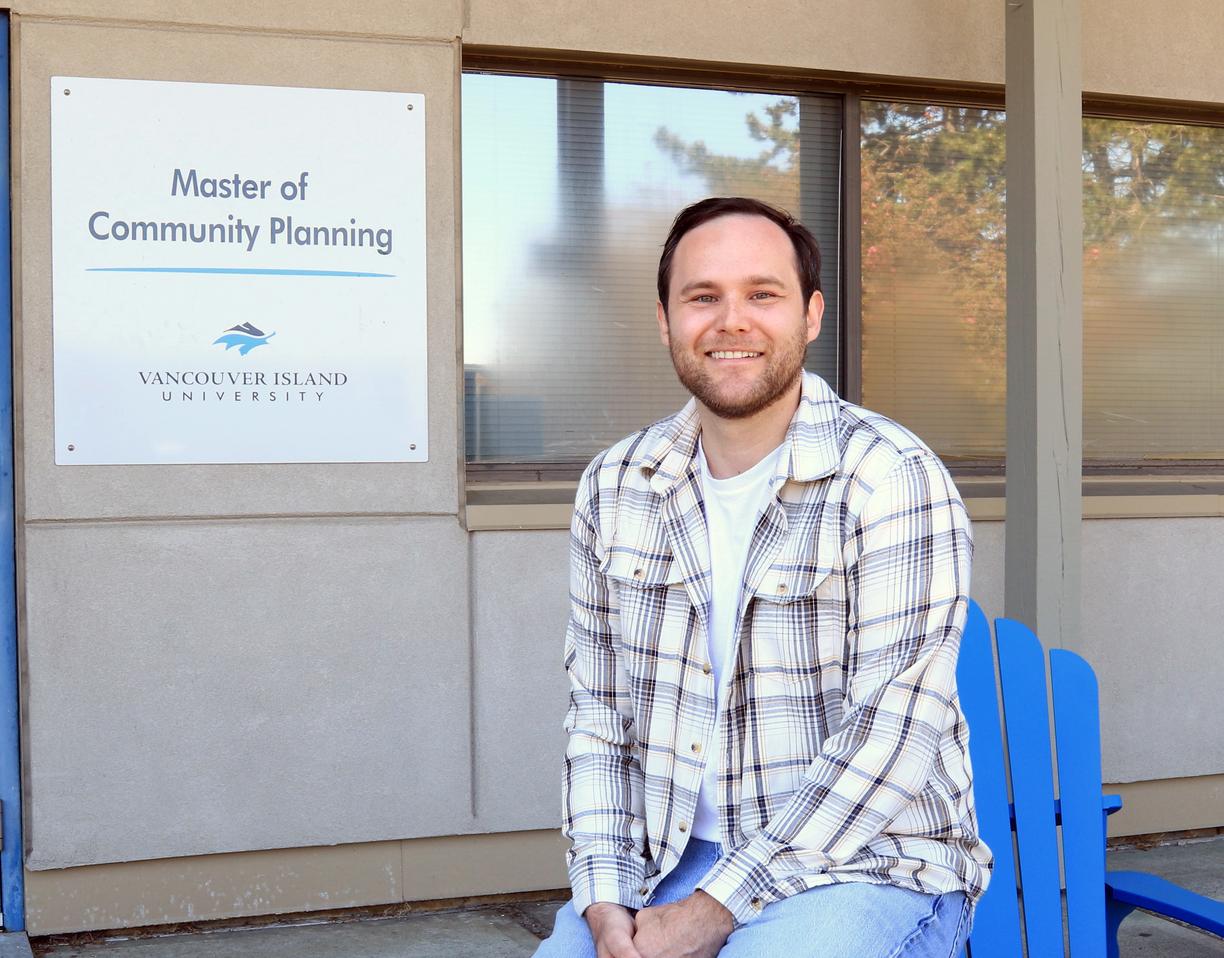 A man wearing a beige and white plaid jacket over a white t-shirt is sitting on the arm of blue Adirondack chair in front of a door and window to a building