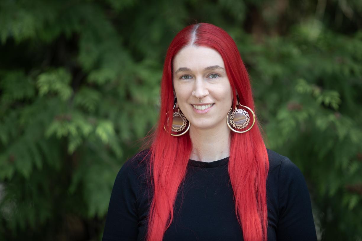 A young woman with bright red, long hair is smiling with tree leaves in the background