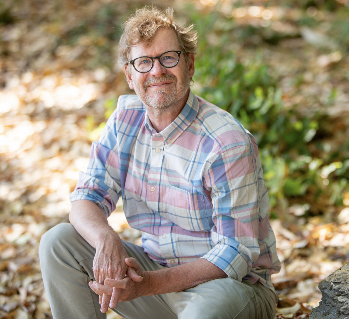 Erik Krogh sitting on the ground outside on a sunny day and smiling at the camera.