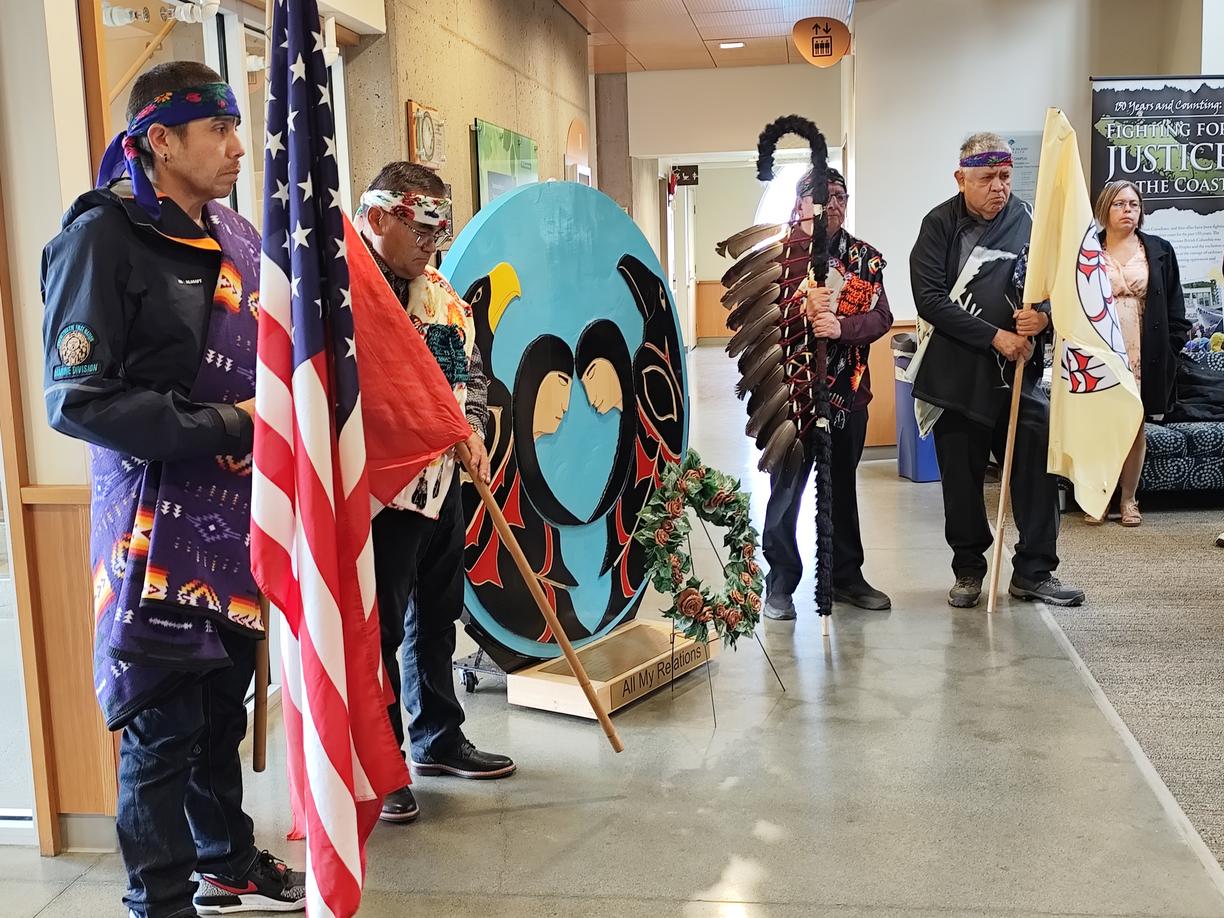 Indigenous community members wearing blankets stand solemnly next to cenotaph next to 