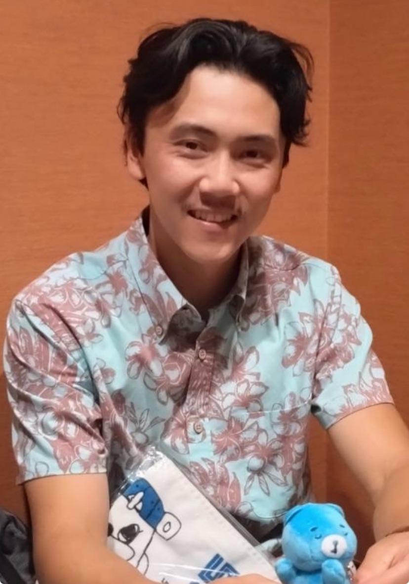 Kenji Boyko smiling. He wears a blue shirt decorated with maroon flowers.