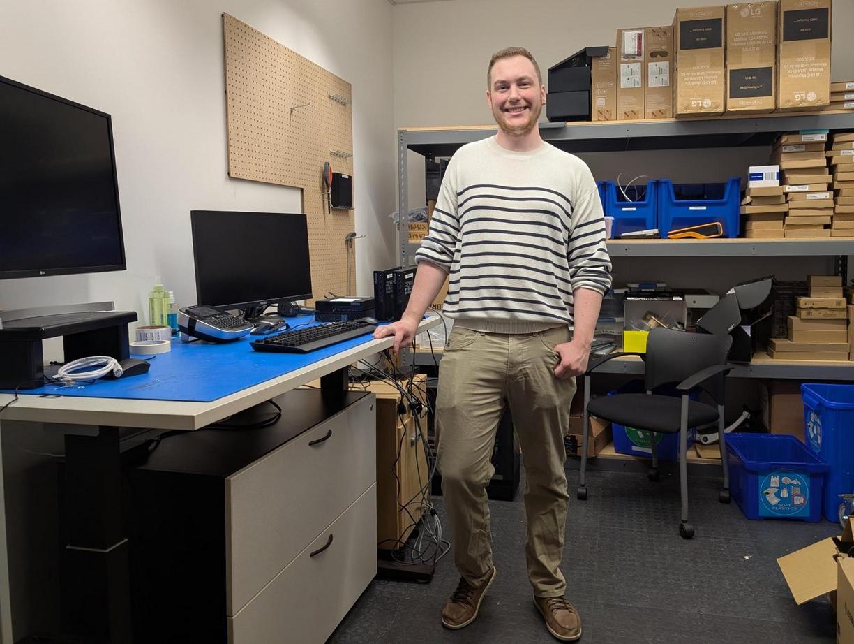 A man standing beside a desk with a computer and a monitor on it in a room with shelves and supplies on them