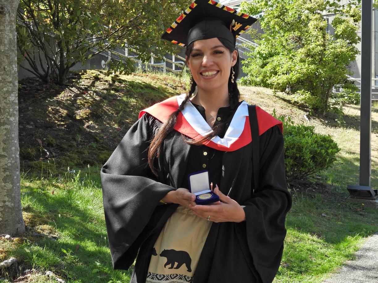 VIU Graduate, wearing convocation robes, Olivia Thomas is holding a medal while standing outside in front of a garden 