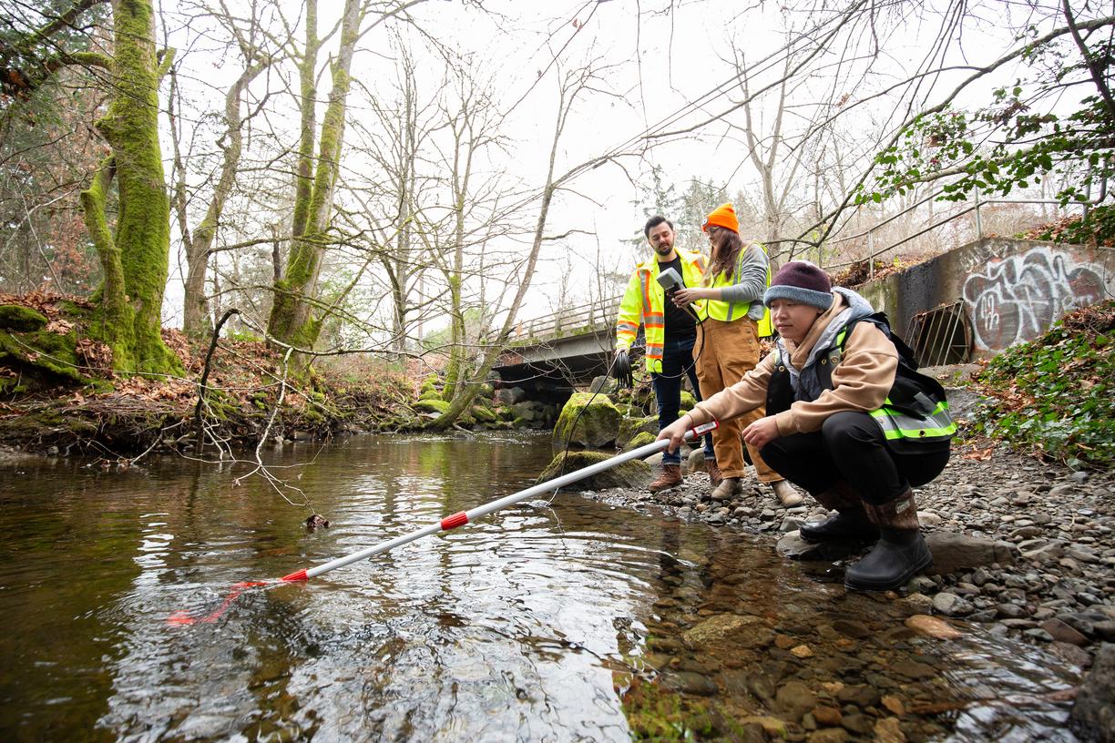 Three people next to a stream, one holds a sampling pole while the others look on