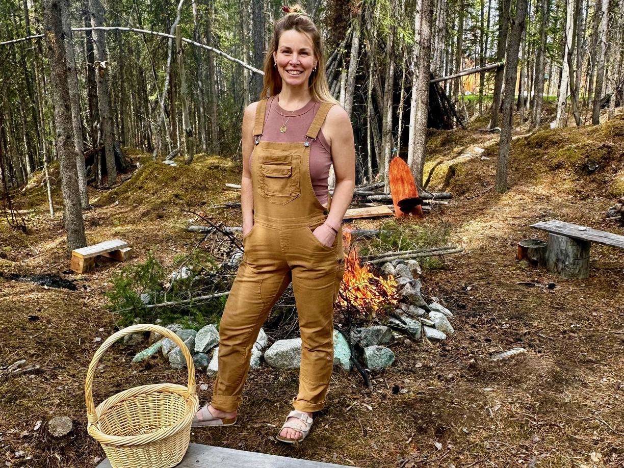 A woman wearing tan overalls is standing in a clearing in the forest in front of a campfire. 