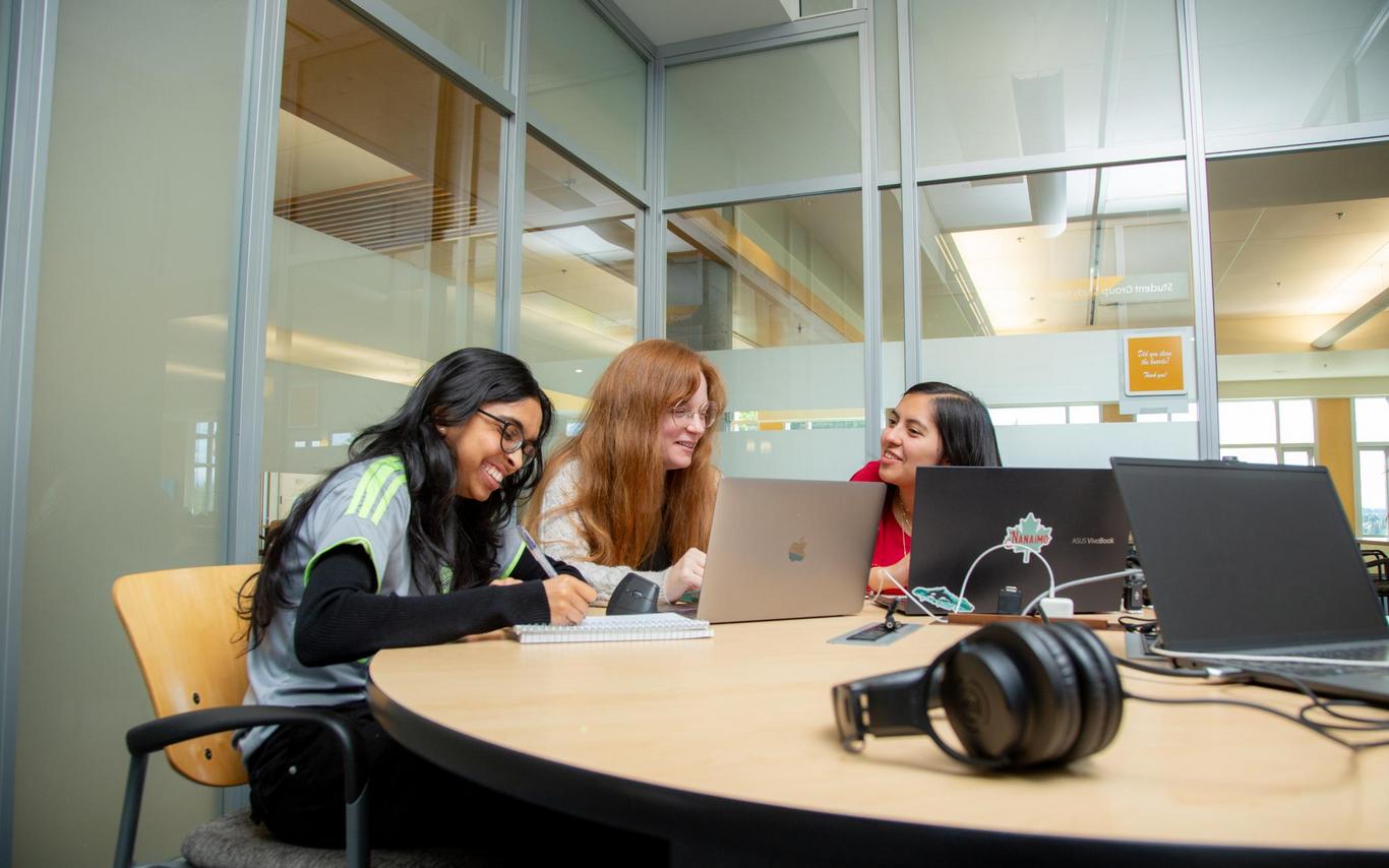 Three students sit at a table and look at a laptop screen together