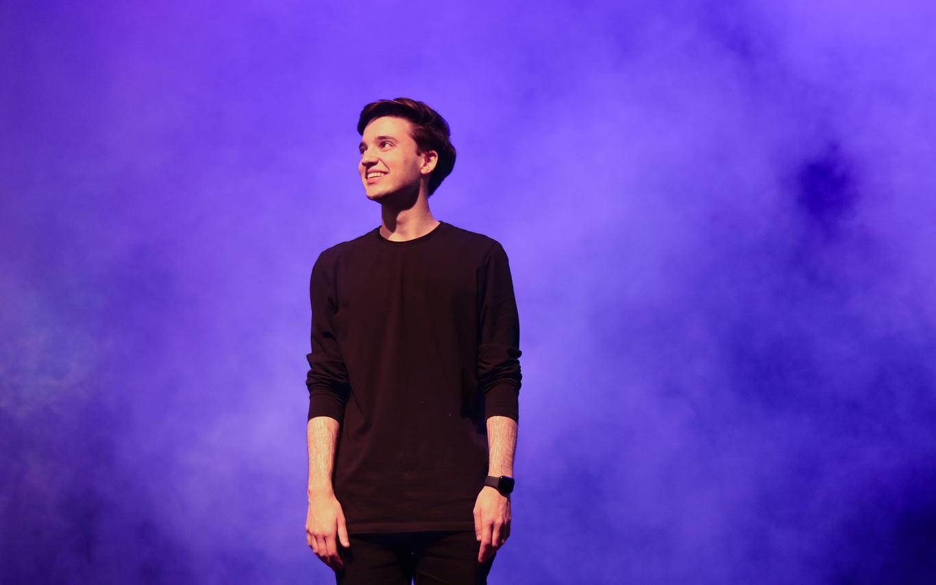 Alex Bhargava stands on the Malaspina Theatre stage looking off to the left. He wears all black and blue and purple smoke swirls around him.
