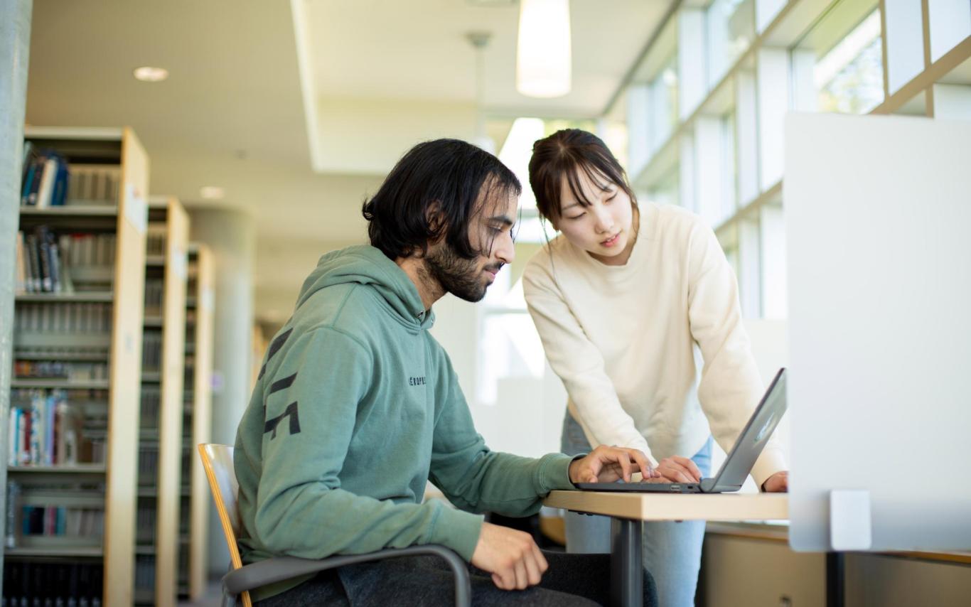 Two people look at a laptop screen in the library