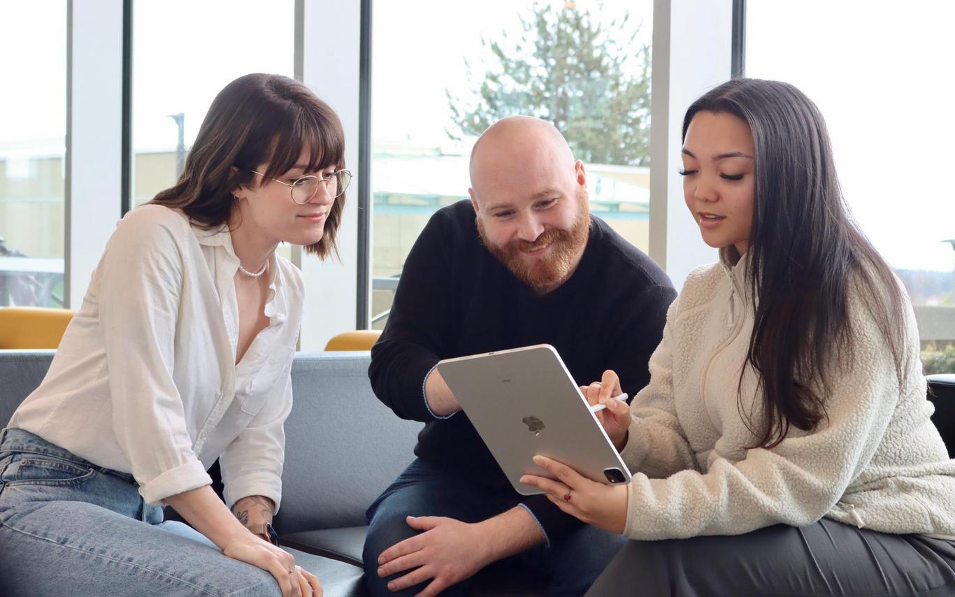 A woman, a man and another woman sitting on a chesterfield looking at a laptop together.