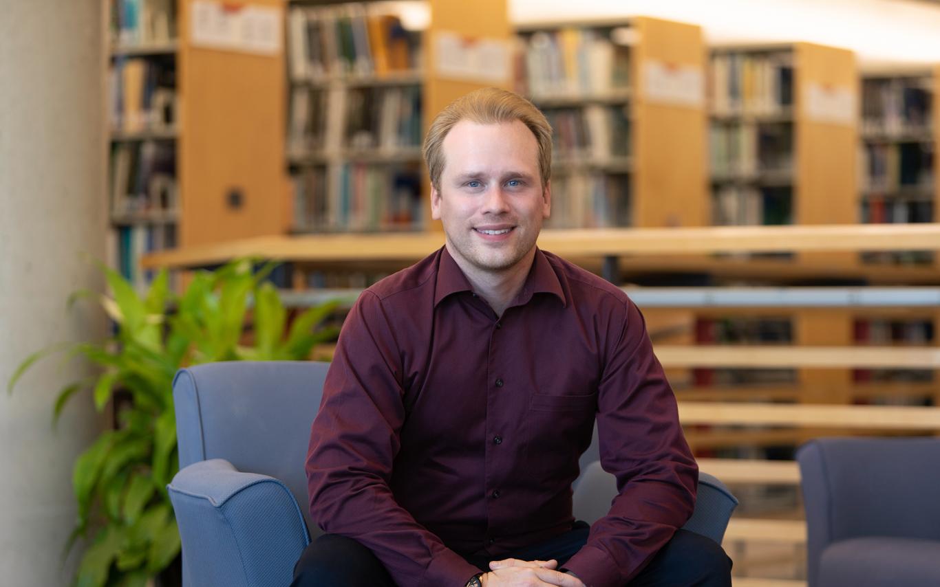 Chris Shanks sits on a blue chair in a library.