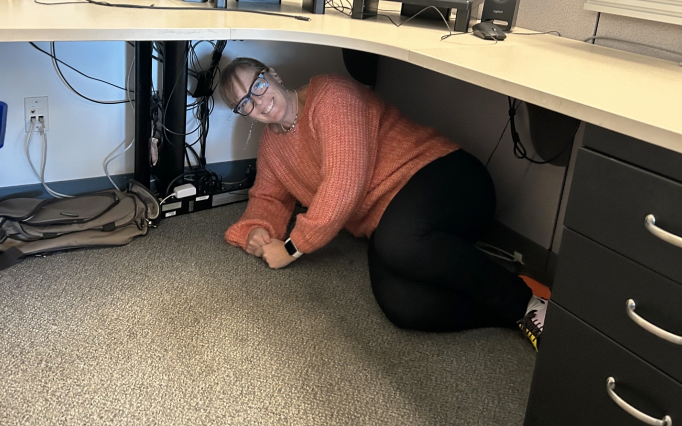 A VIU employee sheltering under a desk and smiling at the camera.