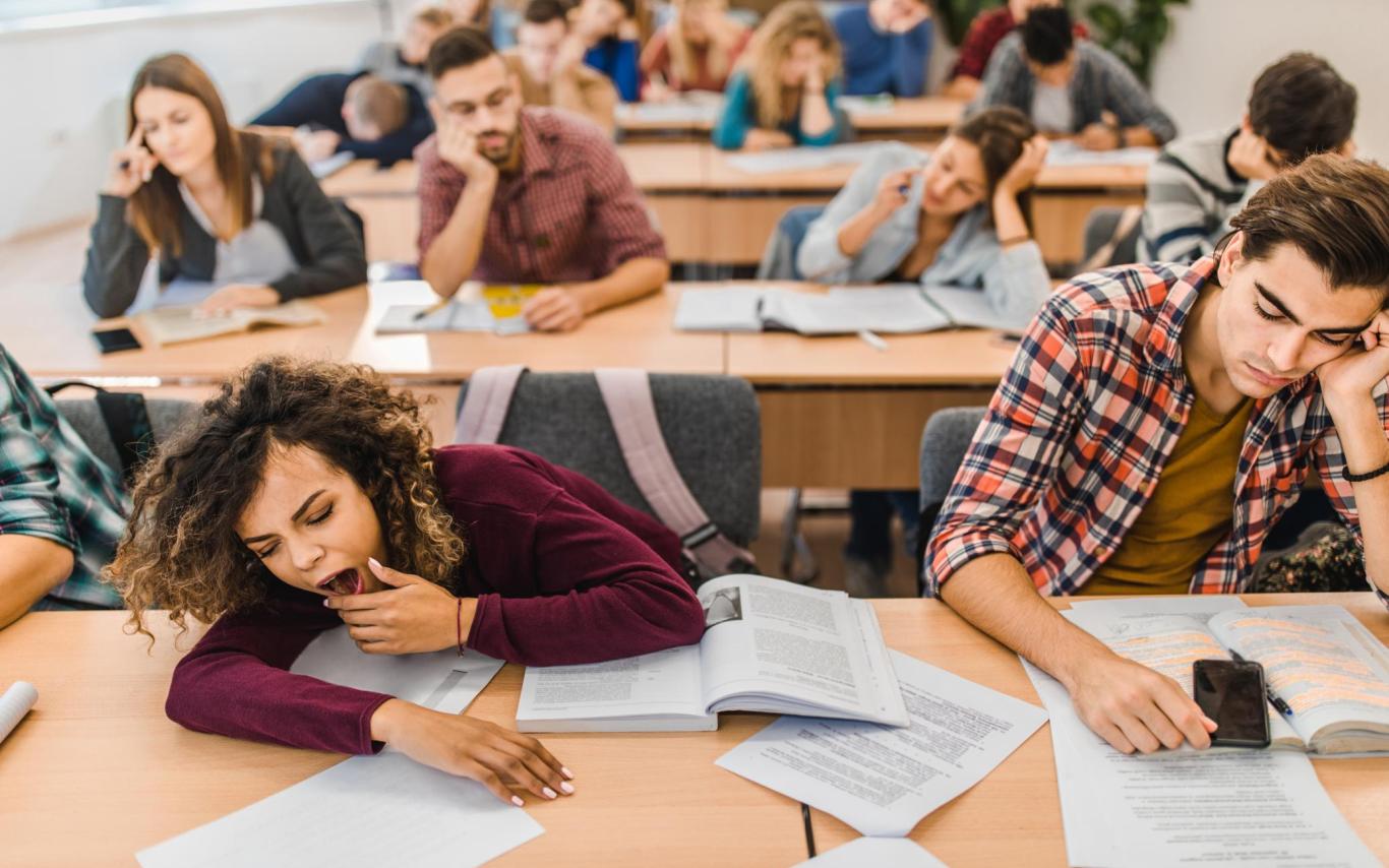 a student on the left yawns while the student next to her has his hand on his face while in a classroom with lots of students in the background