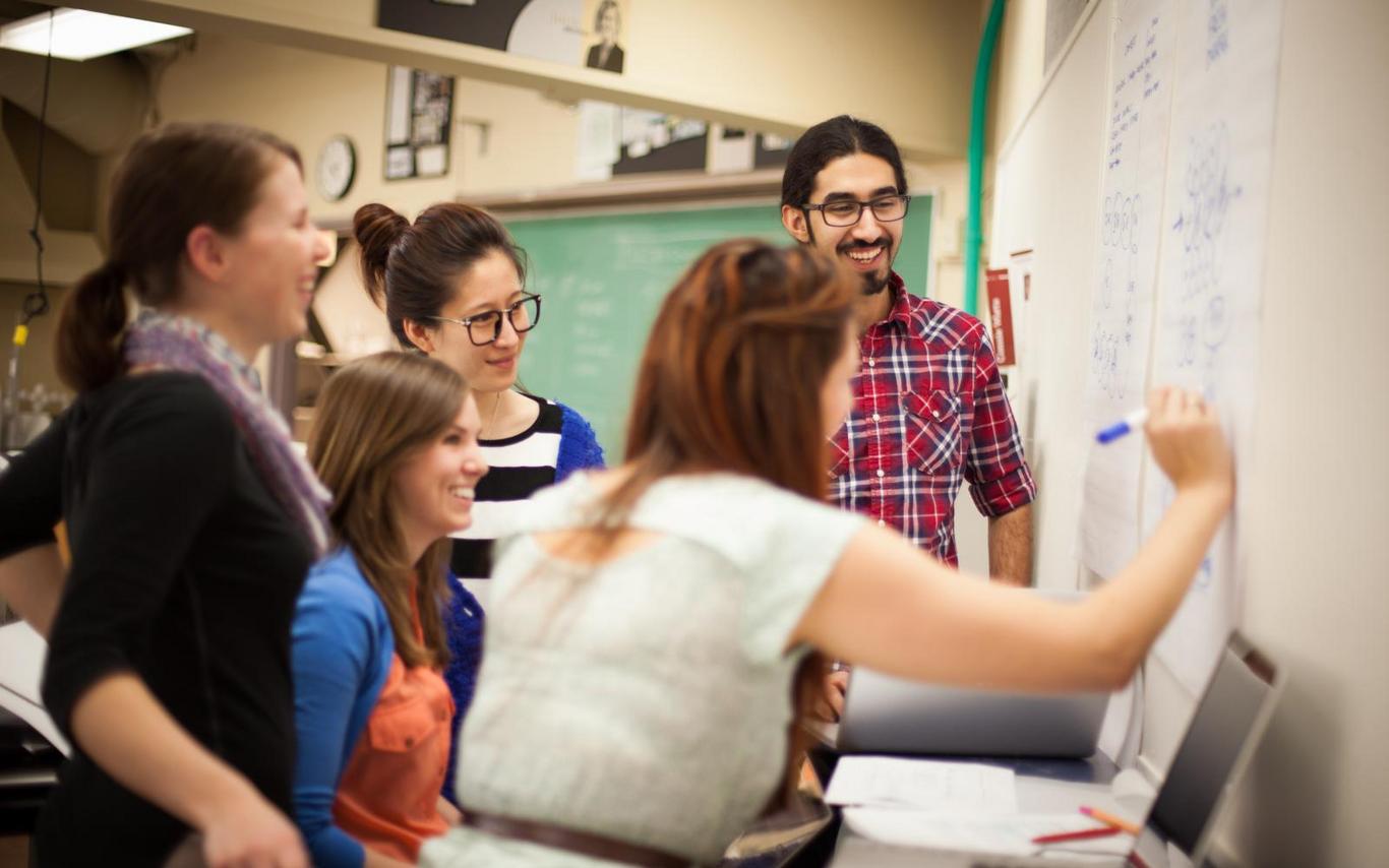 A group of students gathers around a student writing on a chalkboard