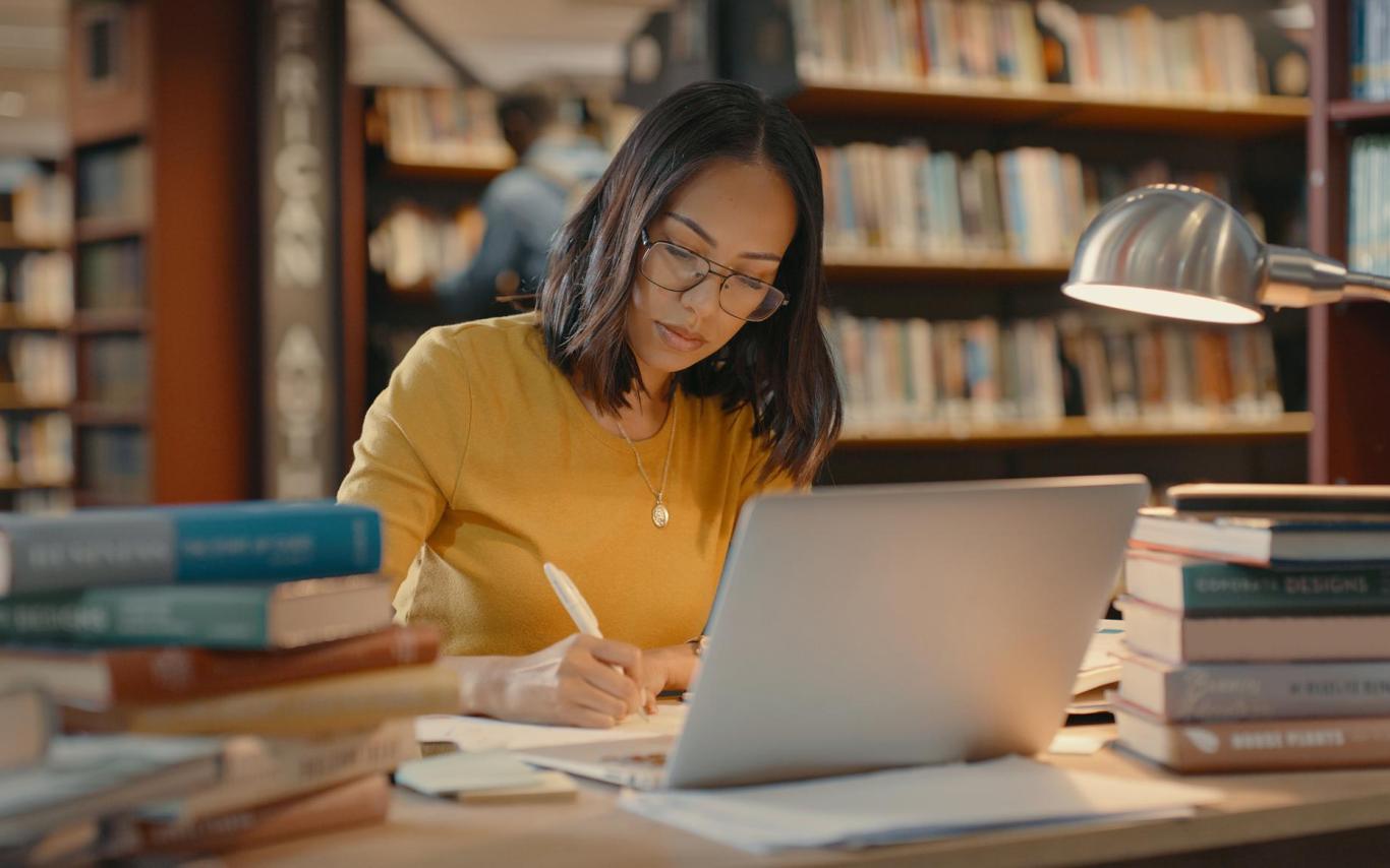 A woman sits at a desk in a library with a laptop open surrounded by books