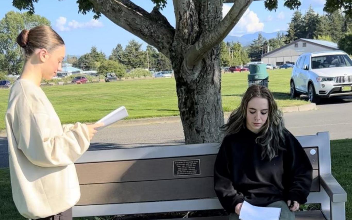Two actors rehearse with scripts in their hand near a bench in a park.