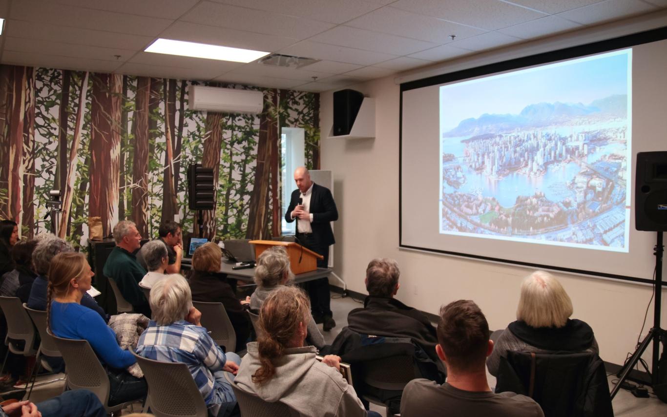 a man speaks next to a screen in front of a packed room