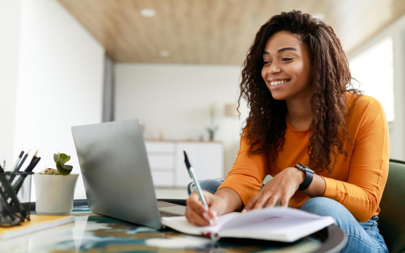 A woman takes notes and smiles while she looks at a laptop screen