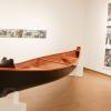 A canoe sits on a stand with drawings hanging on the gallery wall's behind it.