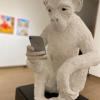An ape made of white plaster and clay holds a cell phone, which it stares at intensely