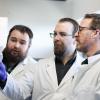 Joseph Nowlan, Scott Britney and Dr. Spencer Russell, wear white lab coats, while looking at a Petri dish.