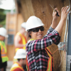 Female electrician wearing. white hard hat a safety vest and sunglasses working on the side of a building and smiling at the camera.
